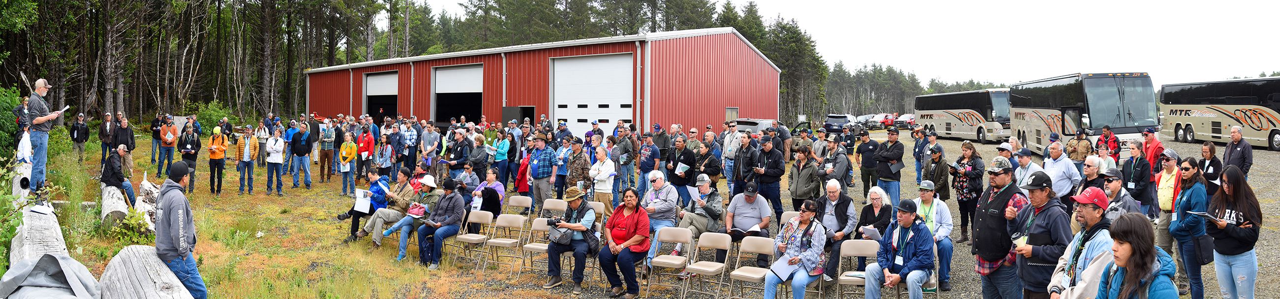 A group of people outside a barn listening to a speaker